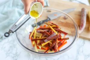 sliced carrots in a glass bowl with oil and herbs being poured on top