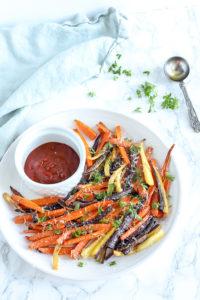 carrot fries topped with fresh herbs and cheese on white place with a small bowl of ketchup